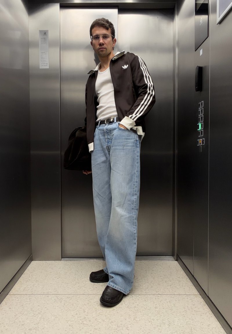 Homme debout dans l'ascenseur portant une veste noire Adidas, un débardeur blanc, un jean bleu clair et des chaussures noires, tenant un sac noir.
