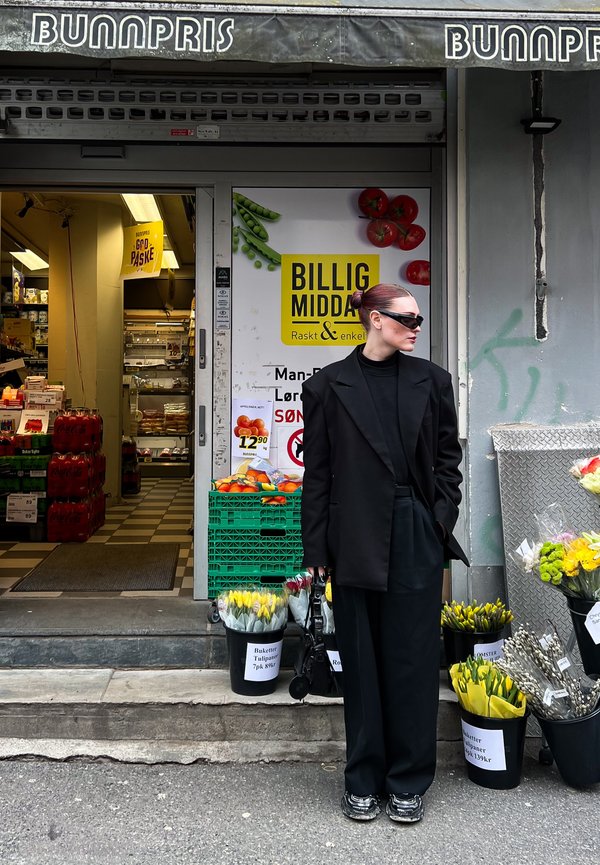 Oversized black blazer worn over a black turtleneck, paired with black wide-leg trousers and sunglasses. Accessory: black handbag. Flower buckets filled with yellow tulips.