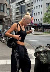 Čierny tielko a voľné čierne tepláky s nápisom "SUPRE" natlačeným po boku. Čierna kabelka s reťazovým popruhom, v ktorej drží šálku kávy.