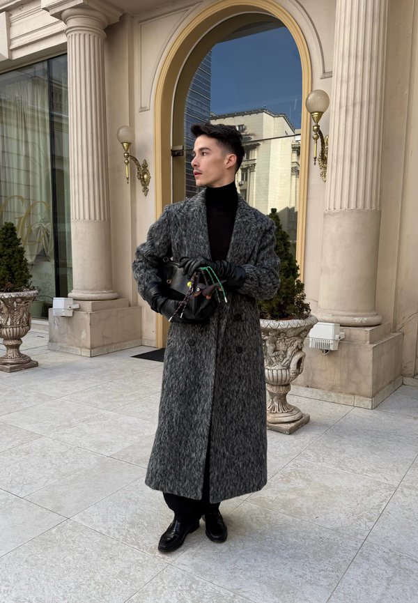 Άνδρας με μακρύ γκρι παλτό και μαύρα γάντια στέκεται κοντά στην είσοδο κτιρίου με αψιδωτή πόρτα και κλασικούς κίονες, κρατώντας μια μαύρη τσάντα.