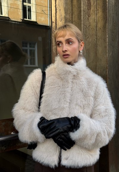 Chaqueta corta de piel sintética en color blanco roto con textura suave, combinada con guantes de cuero negro. Cuenta con un cuello alto y cierre de cremallera.