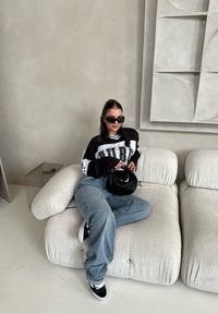 Μαύρη και άσπρη υπερμεγέθης μπλούζα με έντονη γραφή, ανοιχτό μπλε φαρδιά τζιν, μαύρη τσάντα, γυαλιά ηλίου, καθισμένη σε καναπέ από ματ κρέμ υφή.