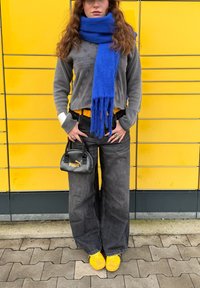 Casaco de fleece cinza, cachecol azul, jeans de pernas largas pretos, cinto amarelo, ténis amarelos e uma pequena bolsa preta com o logótipo da Puma. Fundo amarelo.