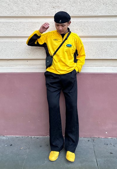 Camiseta deportiva amarilla de mangas largas con detalles negros, pantalones amplios negros y zapatos amarillos. Bolso negro colgado del hombro.