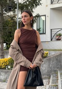 Robe sans manches marron avec des détails drapés, portée sous un trench-coat beige clair. Sac à main noir de forme triangulaire et texture lisse.
