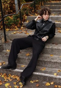 Sort jakke i kunstlæder med grafisk detalje, brede sorte bukser og sorte sko, siddende på stentrapper omgivet af efterårsblade.