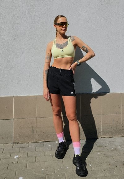 Sujetador deportivo amarillo con logo negro de Adidas, pantalones cortos negros con cordón, zapatillas negras y calcetines ombre en rosa, de pie contra una pared gris.