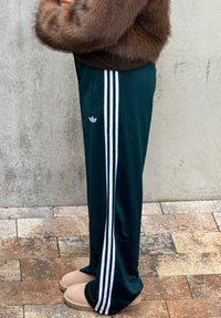 Gröna träningsbyxor med tre vita ränder längs sidorna, med en vit logotyp. Bärs med beige slip-on skor och en brun fluffig tröja.