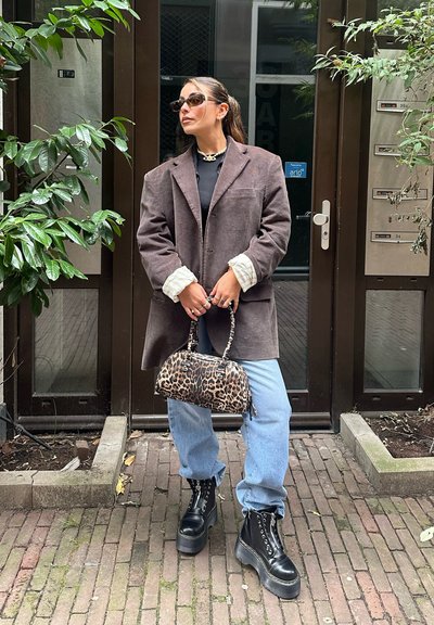 Blazer de pana marrón de gran tamaño, top negro, jeans azul claro, botas negras gruesas de cordones y un bolso de leopardo. Lleva gafas y anillos.