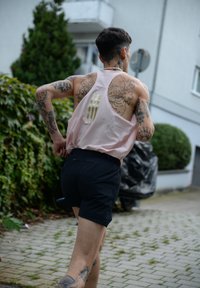 Svijetlo ružičasta bez rukava sportska majica s prugastim detaljem na leđima, uparen s crnim kratkim hlačama. Vidljive tetovaže na rukama i nogama. Vanjsko okruženje s zelenilom.