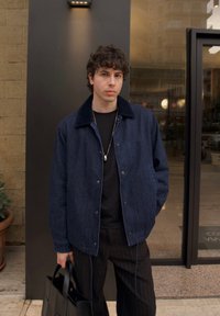 Jaqueta de denim azul escuro com fecho de botão na frente e colarinho preto, combinada com uma t-shirt preta e calças risca de giz, segurando uma bolsa preta.