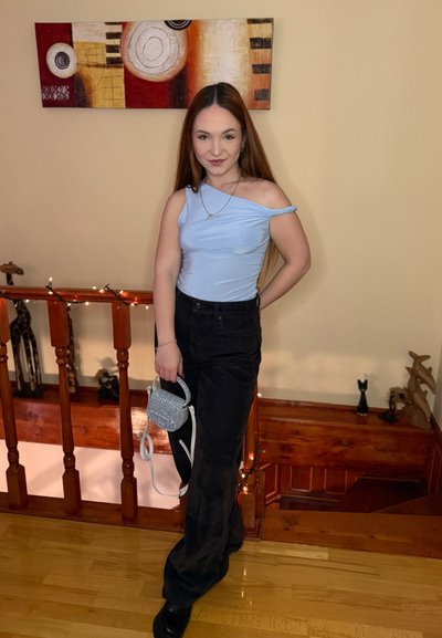 Joven con el cabello largo, vestida con un top azul de un solo hombro y pantalones negros, sosteniendo un pequeño bolso gris, de pie en el interior cerca de una barandilla de madera.