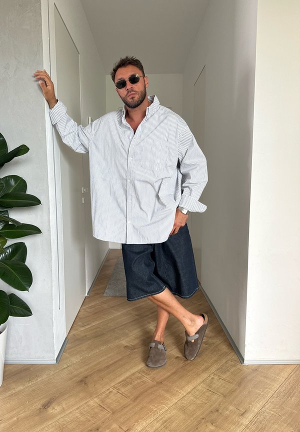 Uomo con occhiali da sole, camicia a righe oversize, pantaloncini in denim e pantofole, in piedi con le gambe incrociate appoggiato a un muro bianco all'interno.