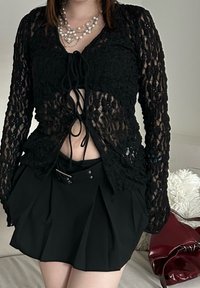O cardigan de renda preta apresenta um design com laço na frente e mangas longas. Combinado com uma saia preta plissada e uma faixa de cintura com detalhes em prata.