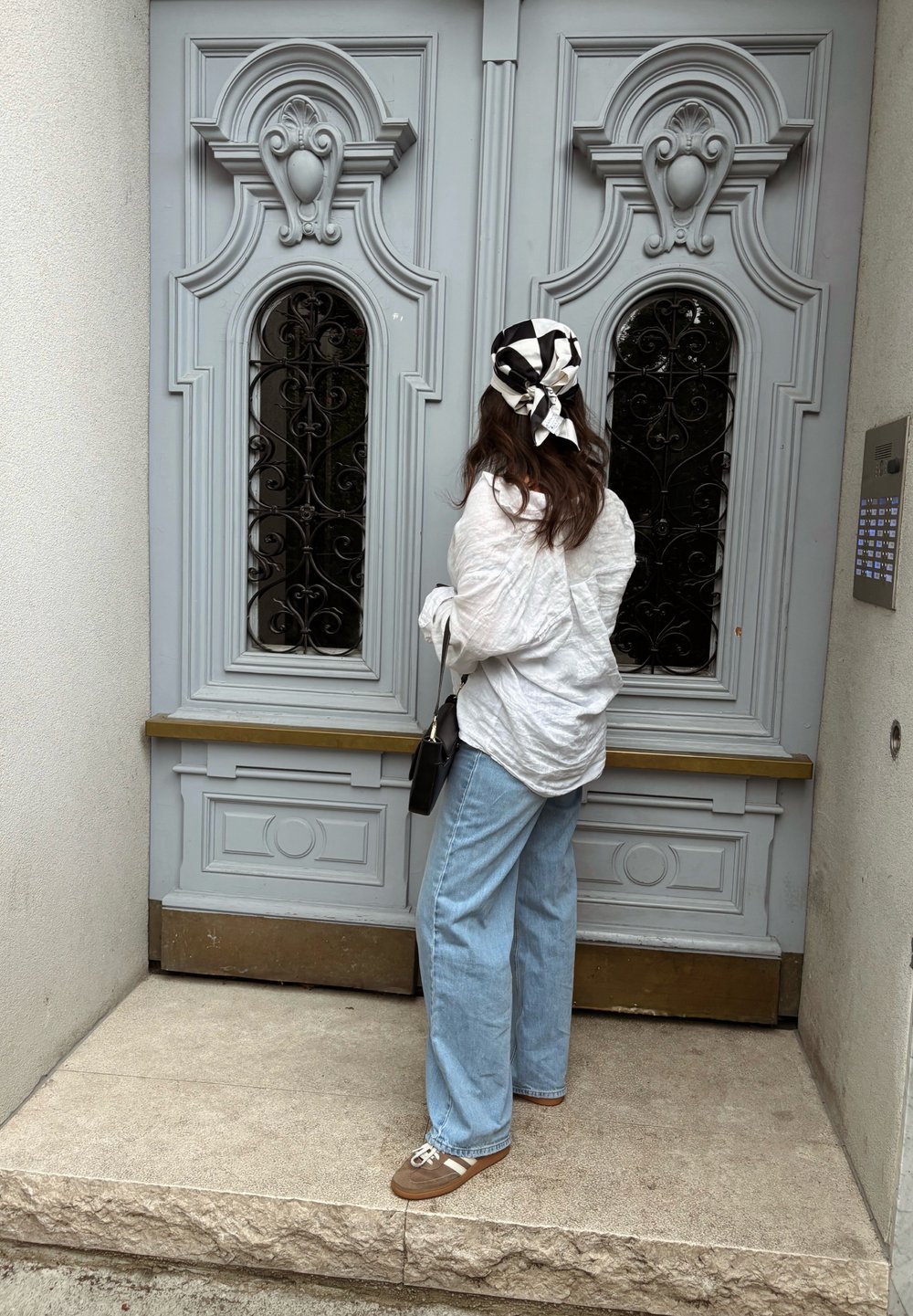 Porta de madeira cinza claro com entalhes ornamentais e grades de metal pretas. Pessoa vestindo uma camisa branca e calças de ganga largas azuis está na entrada.