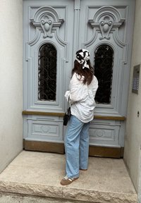 Porta de madeira cinza claro com entalhes ornamentais e grades de metal pretas. Pessoa vestindo uma camisa branca e calças de ganga largas azuis está na entrada.