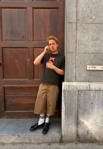 Una persona está de pie contra una puerta de madera, vistiendo una camiseta negra con un gráfico, pantalones cortos caqui y zapatos negros con calcetines blancos a rayas.