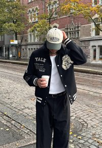 Zwarte varsity jacket met witte strepen en Yale-borduring, met een bulldogafbeelding. Gecombineerd met een off-white pet en zwarte broek.