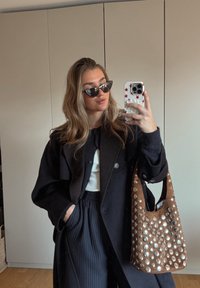 Casaco oversized preto, blusa branca, calças risca de giz, óculos de sol em forma de olho de gato. Bolsa castanha com tachas metálicas. Reflexo no espelho captura o conjunto completo.