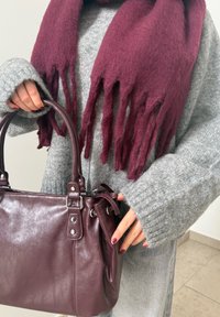 Personne portant un pull en maille gris et une écharpe bordeaux, tenant un sac à main en cuir bordeaux assorti à son vernis à ongles.