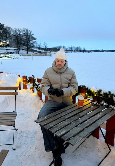 Hombre con un gorro blanco y chaqueta beige sentado en una mesa al aire libre nevada con una barandilla decorada y una bebida caliente cubierta con crema batida.