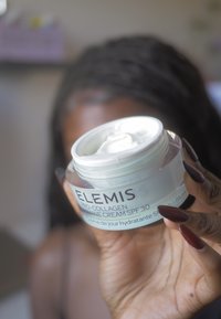 Una mano con lunghe unghie scure che tiene aperto un barattolo di Elemis Pro-Collagen Marine Cream SPF 30, con una persona sfocata sullo sfondo.