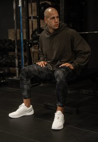 Conjunto atlético composto por um hoodie oversized verde escuro, leggings pretas texturizadas com padrão e sapatilhas atléticas brancas.