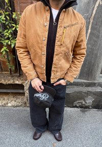 Uomo che indossa una giacca beige sopra un maglione nero e jeans scuri, che tiene un berretto nero con scritta bianca, in piedi su un pavimento di pietra vicino a un muro con piante.