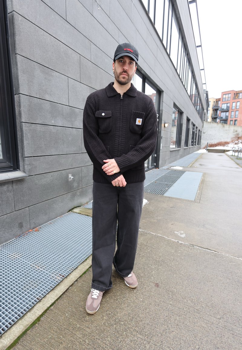 Homme en veste noire en tricot, pantalon gris et casquette, debout sur un trottoir urbain à côté d'un bâtiment moderne gris par une journée nuageuse.