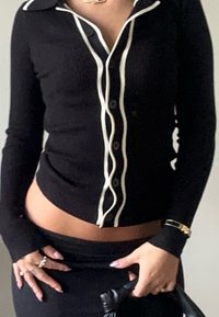 Top de manga longa preto com maxi-rib, apresentando um acabamento em branco, um decote com gola e fecho de botão; combinado com uma saia preta e acessórios.