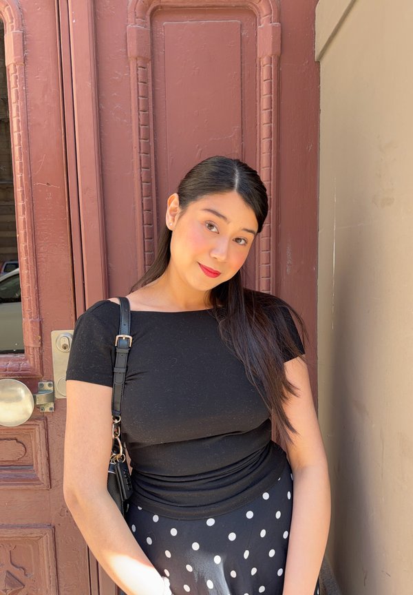 Jonge vrouw met lang donker haar, die een zwart topje en een zwarte rok met witte stippen draagt, staat tegen een bruine deur en draagt een zwarte tas.