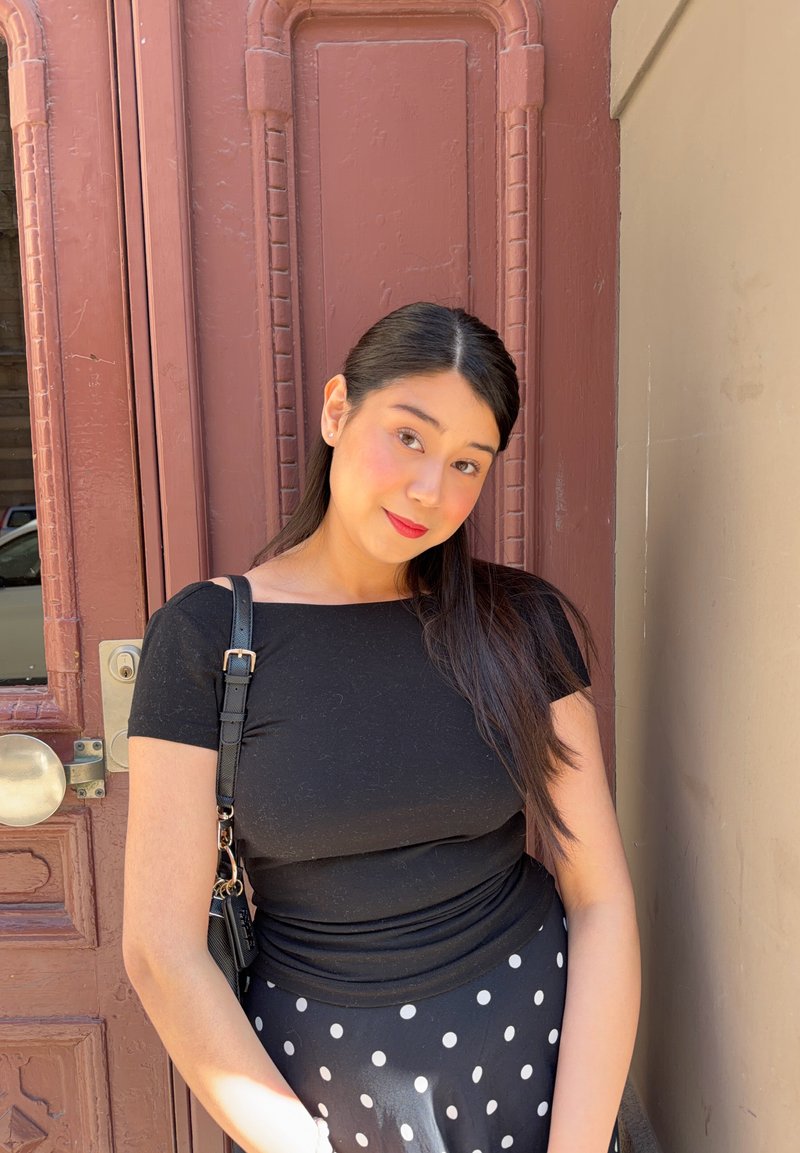 Jeune femme aux longs cheveux foncés portant un haut noir et une jupe noire à pois blancs, debout contre une porte marron, portant un sac noir.