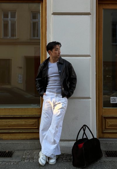 Chaqueta de cuero negra, camisa gris ajustada, pantalones blancos de pierna ancha y zapatillas grises. Bolsa de mano negra con interior rojo en el suelo cerca.