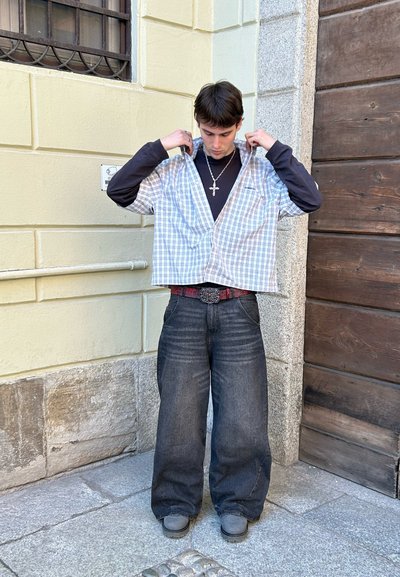 Camisa de cuadros de manga corta en azul claro y blanco, superpuesta a una camiseta de manga larga negra, combinada con jeans de mezclilla oscuros de corte holgado.