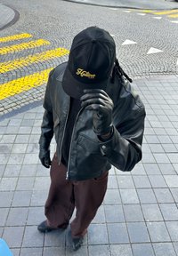 Personne portant une veste en cuir noire, des gants noirs, une casquette noire avec texte jaune, et un pantalon marron debout sur une rue pavée avec des lignes de passage piéton jaunes.