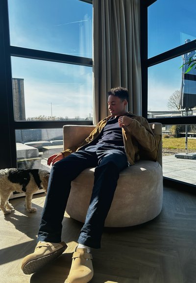 Silla circular de terciopelo beige con una textura suave, una persona que lleva una chaqueta marrón y pantalones negros, y un pequeño perro cerca sobre un suelo de madera.