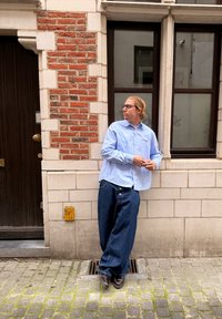 Vaaleansininen nappipaita yhdistettynä löysiin tummansinisiin farkkuihin. Seisoo karkeaa tiiliseinää vasten, taustalla puinen ovi.