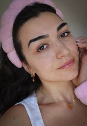 Diadema de tela rosa, pendientes de oro con acentos circulares, tono de piel claro, labios brillantes y un calentador de brazos rosa texturizado. Luz natural.