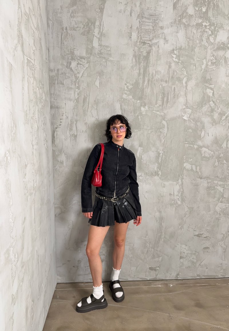 Personne portant une veste noire et une jupe plissée, des chaussettes blanches, des sandales noires et un sac à main rouge, debout contre un mur gris texturé.