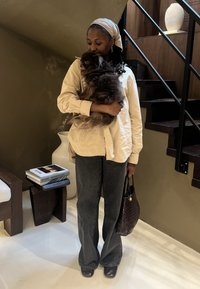 Beige skjorte med knapper, mørke utvidede jeans, flettet veske, holder en brun fluffy hund. Bakgrunnen har en teksturert vegg og minimalistisk innredning.