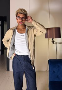 Άτομο με ξανθά σγουρά μαλλιά και γυαλιά ποζάρει φορώντας μπεζ σακάκι, λευκή μπλούζα Versace και σκούρο μπλε παντελόνι σε έναν σύγχρονο χώρο.