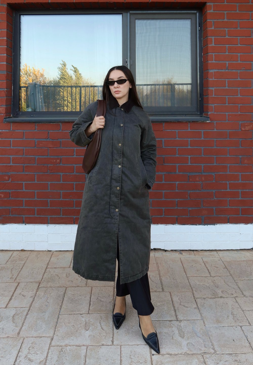 Donna con cappotto lungo scuro, occhiali da sole neri, che porta una borsa marrone, in piedi su un pavimento piastrellato davanti a un muro di mattoni rossi con una grande finestra.