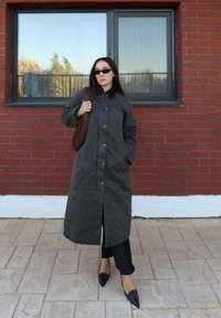 Donna con cappotto lungo scuro, occhiali da sole neri, che porta una borsa marrone, in piedi su un pavimento piastrellato davanti a un muro di mattoni rossi con una grande finestra.