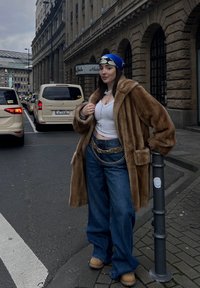 Καφέ ψεύτικη γούνινη καμπαρντίνα, λευκό κοντό τοπ, μπλε παντελόνια φαρδιάς γραμμής, στρωτές αλυσίδες στη μέση, μπεζ μπότες, μπλε και λευκή μοτίβο κορδέλα για τα μαλλιά.