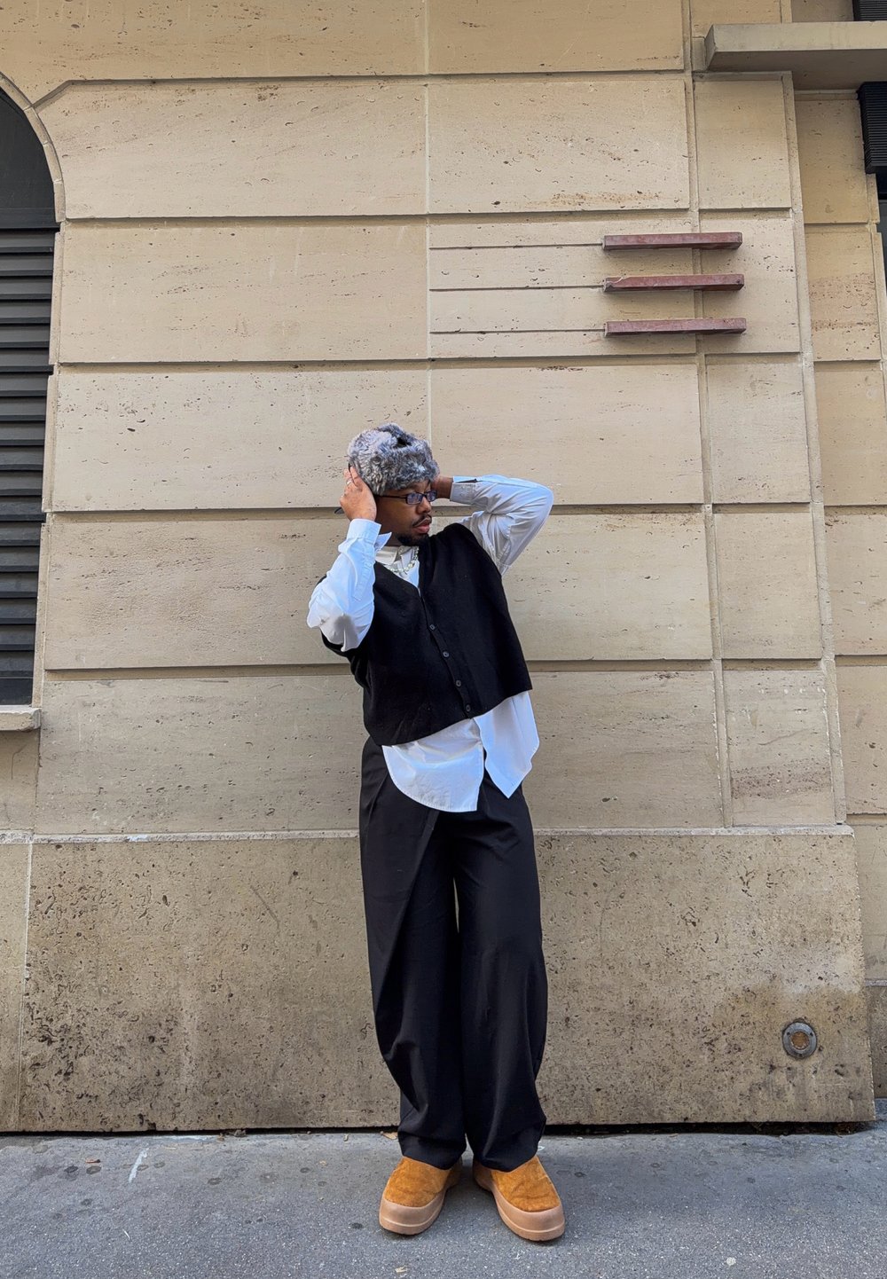 Homme portant un chapeau en fourrure grise, des lunettes, un gilet noir, une chemise blanche, un pantalon noir large et des chaussures beige, debout contre un mur en pierre beige, ajustant son chapeau.