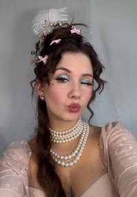 Una collana di perle a strati, un vestito rosa chiaro senza spalle, capelli ricci adornati da un accessorio per capelli floreale e di piume, e ombretto azzurro chiaro.