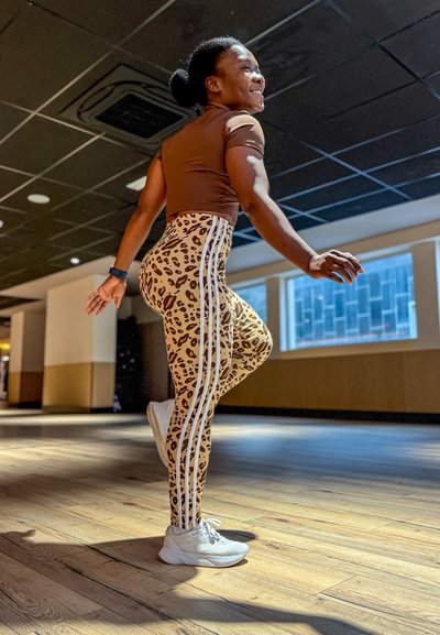 Top marrón ajustado, leggings de estampado de leopardo con rayas blancas, zapatillas blancas. Suelo de madera y ventanas brillantes al fondo.