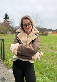 Brun shearlingjakke med beige pelskrave og manchetter, med lynlåslukning og sorte detaljer. Båret over en let sweater og sorte bukser.