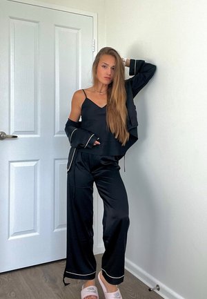 Conjunto de pijama de satén negro con ribete dorado, que incluye un top sin mangas y pantalones de pierna ancha. Combinado con sandalias tipo slide de color claro.