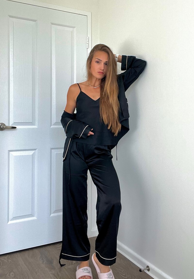 Schwarzes Satin-Pyjama-Set mit goldenen Paspelierungen, bestehend aus einem ärmellosen Oberteil und weiten Hosen. Kombiniert mit hellen Pantoletten.